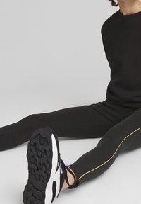 Leggings sportivi neri con strisce laterali dorate, abbinati a sneakers bianche e nere con suole testurizzate. Il tessuto appare liscio ed elastico.