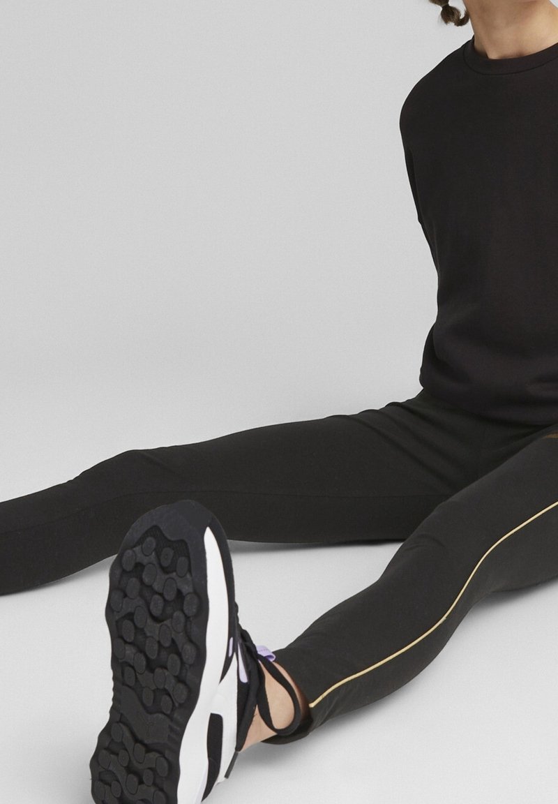 Leggings sportivi neri con strisce laterali dorate, abbinati a sneakers bianche e nere con suole testurizzate. Il tessuto appare liscio ed elastico.