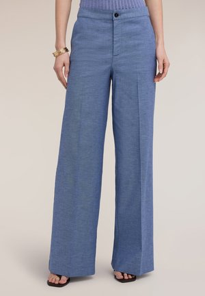 Femme portant un pantalon bleu taille haute à jambes larges avec une fermeture à boutons, associé à des sandales noires à bout ouvert et des bracelets dorés au poignet gauche.