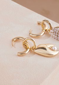 Örhängen i guldfärg med cirkulära ringar och en kloliknande design med en liten pärla som detalj. Slät, glänsande yta och noggrant hantverk.