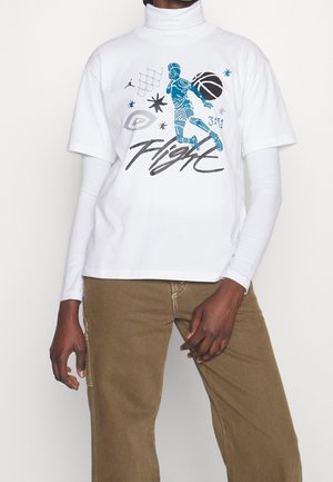 Personne portant un haut blanc à couches avec un graphisme de joueur de basket-ball, des étoiles et le texte "Flight", accompagné d'un pantalon marron.