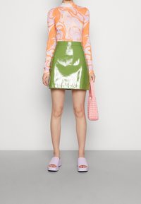 Frau in einem grünen, glänzenden Minirock, orange- und pinkfarbenem Langarmoberteil, mit einer pinken Handtasche und lilafarbenen Plateauschuhen.