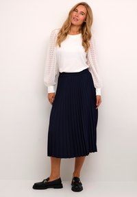 Vit topp med texturerade puffärmar, i kombination med en knälång marinblå plisserad kjol; komplett med svarta loafers med gulddetaljer.