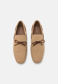 Tan suede loafers with brown laces, featuring a textured upper, gold-tone accents, and a cushioned insole for comfort.