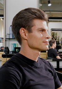 Mannelijke model met kort, glad bruin haar zit in een salon. Hij draagt een zwart V-hals t-shirt. De achtergrond bevat salonproducten en spiegels.