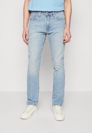 Personne portant un jean bleu clair avec une petite déchirure sur la cuisse gauche, des baskets blanches et une chemise bleu clair, debout devant un fond uni.
