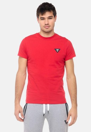 Uomo in piedi di fronte, indossa una maglietta rossa a maniche corte e pantaloni della tuta grigio chiaro con coulisse e dettagli con zip nere.