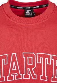 Roter Sweatshirt mit geripptem Rundhalsausschnitt und gesticktem "STARTER"-Logo in Weiß auf der Vorderseite. Der Stoff erscheint glatt mit einer leichten Struktur.