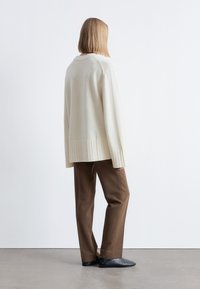 Hellbeige übergroßer Pullover mit geripptem Saum, kombiniert mit braunen, maßgeschneiderten Hosen und schwarzen flachen Schuhen, von hinten gesehen vor einem schlichten Hintergrund.