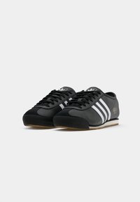 Pár černých tenisek Adidas s bílými pruhy, černými tkaničkami a gumovými podrážkami, předložených na jednobarevném bílém pozadí.