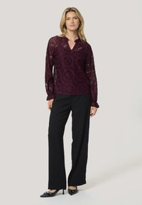 Blusa di pizzo bordeaux con intricati motivi floreali, maniche lunghe e scollatura a V, abbinata a pantaloni neri a gamba larga e décolleté a punta.