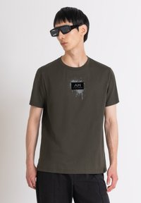 Camiseta de algodón verde oliva con mangas cortas, que presenta un logotipo rectangular negro con detalles de salpicaduras. Se lleva con gafas de sol negras de estilo angular.