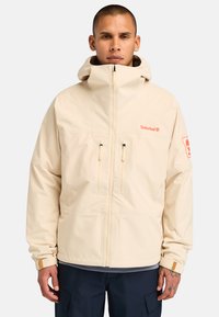 Beige vedenkestävä takki, jossa on korkea kaulus, kaksi etuvetoketjua ja logo rinnassa. Ominaisuuksiin kuuluvat säädettävät hihansuut ja huppu.