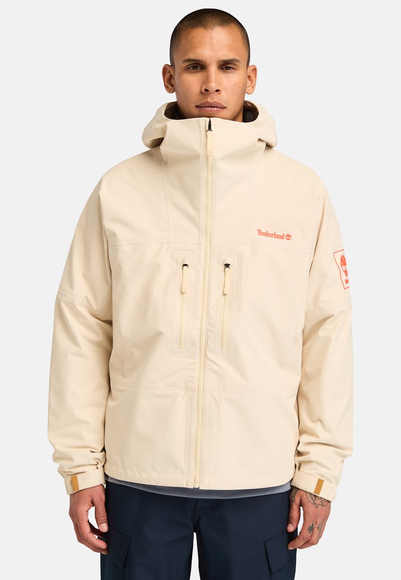 Beige vedenkestävä takki, jossa on korkea kaulus, kaksi etuvetoketjua ja logo rinnassa. Ominaisuuksiin kuuluvat säädettävät hihansuut ja huppu.