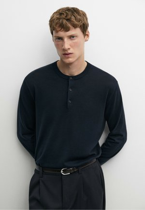 Junger Mann mit heller Haut und kurzen lockigen Haaren trägt ein schwarzes Henley-Langarmshirt und schwarze Hose vor weißem Hintergrund.