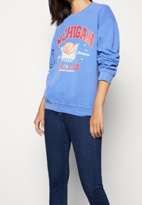 Sudadera azul claro con cuello redondo que presenta un gráfico de baloncesto y texto rojo que dice "MICHIGAN" y "CLUB DEPORTIVO."
