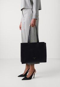 Svart skinn tote bag med strukturert overflate, to lange håndtak og gull logo detalj foran. Kombinert med et rutete antrekk og høyhælte sko.
