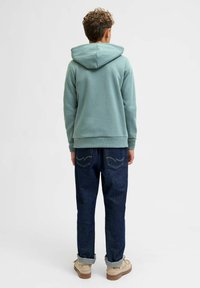 Sudadera verde con cordón ajustable, material de algodón, corte relajado. Combinada con jeans de mezclilla azul oscuro y zapatos beige con los puños remangados.