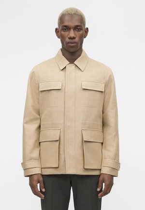 Mand med kort blondt hår iført en beige jakke med fire klaplommer og sorte bukser, stående foran en hvid baggrund.