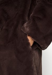 Bruine mantel van namaakbont met een zachte, pluizige textuur; voorzien van een zichtbare zijzak en wijde mouwen.