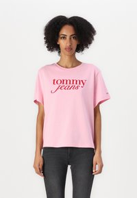 Rosa bomulls t-skjorte med korte ermer, med en stor rød "tommy jeans" logo over brystet. Avslappet passform, glatt tekstur.