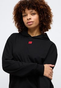 Fekete kapucnis pulóver, puha anyagból készült, kis piros logóval a mellrészen és hosszú ujjak fitted mandzsettával.