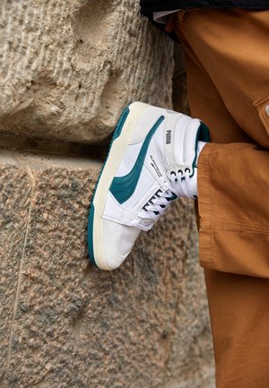 Osoba nosząca białe i turkusowe wysokie sneakersy Puma z białymi sznurowadłami i brązowe spodnie, stopa oparta na fakturowanej kamiennej ścianie.