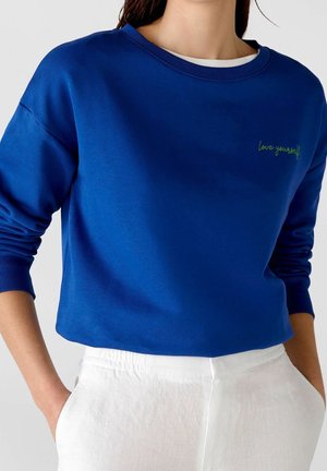 Primo piano di una persona che indossa un felpa blu con la scritta "ama te stesso" ricamata sul petto e pantaloni bianchi con le mani nelle tasche.