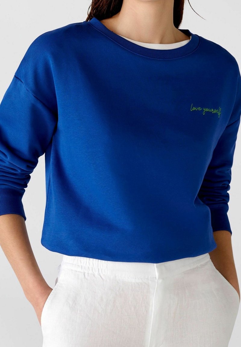 Primer plano de una persona que lleva un sudadera azul con "amáte a ti mismo" bordado en el pecho y pantalones blancos con las manos en los bolsillos.