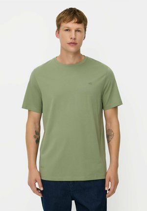 Mann mit kurzen hellbraunen Haaren, der ein einfarbiges hellgrünes kurzärmeliges T-Shirt und dunkle Hosen trägt, steht vor einem weißen Hintergrund.