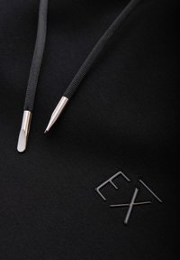 Schwarzer Stoff mit zwei schwarzen Kordeln mit silbernen Metallspitzen und erhabenen schwarzen Buchstaben "EXI" auf der Oberfläche.