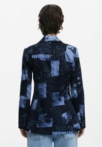Blazer marine avec un design en patchwork, featuring des impressions photographiques bleues et un texte manuscrit. Coupe ajustée avec des revers notched et des manches longues.