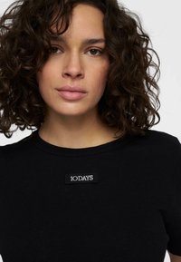 Zwarte katoenen t-shirt met ronde hals, met een klein wit "10 DAYS" logo patch op de voorkant gestikt. Glad textuur.