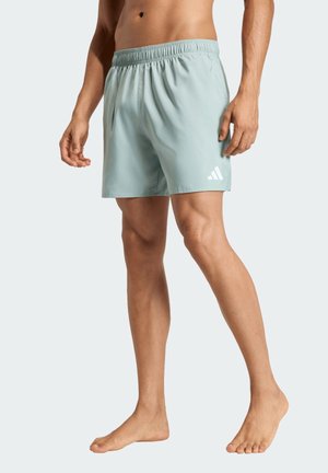 Mężczyzna w jasnoniebieskich sportowych szortach Adidas, stojący boso na jednolitym jasnym tle.