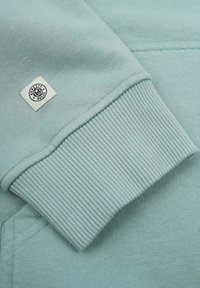 Mintgrön sweatshirt med ribbad mudd och krage. Har en fyrkantig etikett med texten "MODERN RULES" sydd på ärmen. Slät textur.