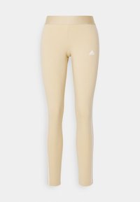 Beige leggings gjorda av elastiskt tyg, med en hög midja och vita sidostripes. Adidas-logotypen är placerad på vänster lår.