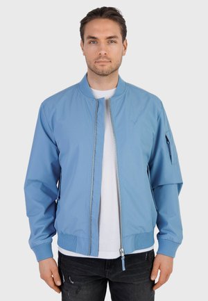 Mężczyzna ubrany w jasnoniebieską bomberkę na zamek błyskawiczny, pod którą ma białą koszulę, oraz czarne, przetarte dżinsy, stoi na tle gładkiego, szarego tła.