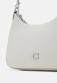Sac à main en cuir blanc texturé avec fermeture éclair argentée, logo "C" argenté et fermoir en métal attachant la bandoulière.