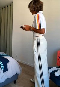 Γυναίκα με λευκή φόρμα Adidas στέκεται μέσα σε εσωτερικό χώρο και κοιτάζει ένα τηλέφωνο, με κρεβάτι και ρούχα ορατά στο παρασκήνιο.