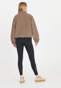 Brauner Fleece-Pullover mit hohem Kragen, oversize Fit und Seitentaschen, kombiniert mit schwarzen Leggings und beigen Sneakers.