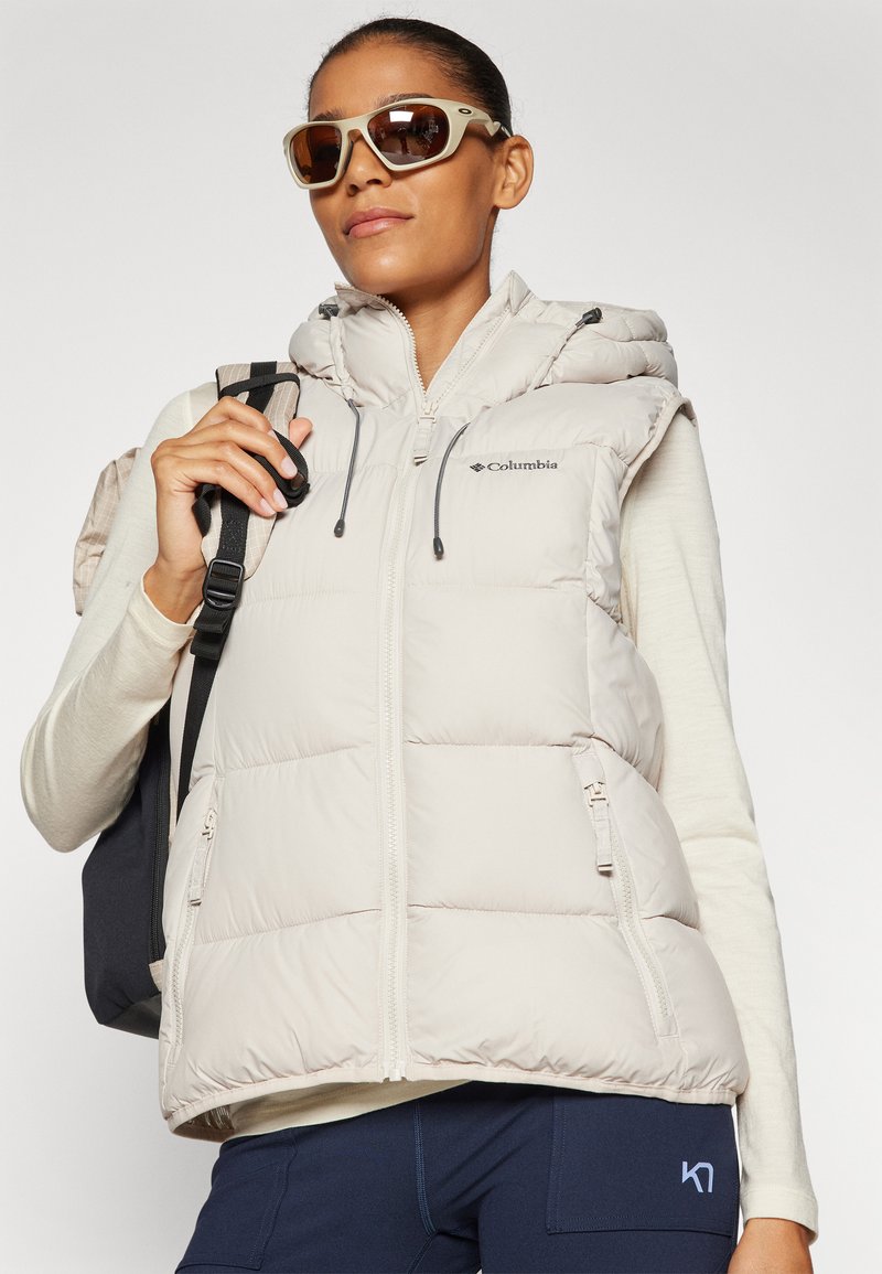 Lichtbeige puffervest met een capuchon, ritssluiting aan de voorkant en verstelbare trekkoorden; gecombineerd met een lichte top en donkere leggings.