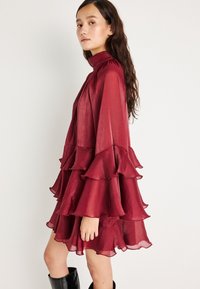 Bordeaux kjole med volanger, høy krage og lange ermer. Fint teksturert, lett stoff. Båret med svarte støvler til kneet.