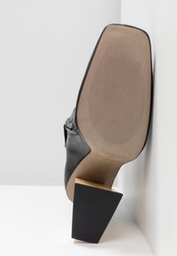 Chaussure à talon en cuir noir posée sur son talon, montrant une semelle lisse de couleur beige clair avec une texture antidérapante légère et un talon bloc sur une surface blanche.