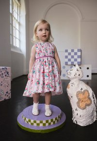 Gėlėta rožinė suknelė su susmulkintu liemeniu ir baltais sandalais; vaikas stovi ant violetinės pakylos šalia raštuoto kartoninio katės skulptūros.