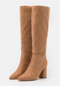 Botas de tacón de ante color trigo hasta la rodilla, con punta afilada, tacón cuadrado y textura suave; líneas limpias y acentos mínimos.