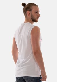 White sleeveless t-shirt with a smooth texture and a round neck, featuring a small logo at the lower left hem, paired with blue denim jeans.