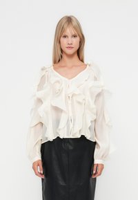 Hvit, volangbluse med lange ermer og rund halsing, med knapper foran. Båret over et svart skinnskjørt.