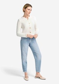 Cardigan en tricot blanc texturé avec un col rond et fermeture par boutons, associé à un jean bleu clair et des ballerines beiges.