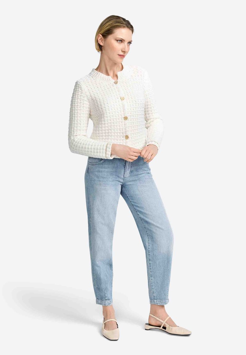 Cardigan en tricot blanc texturé avec un col rond et fermeture par boutons, associé à un jean bleu clair et des ballerines beiges.