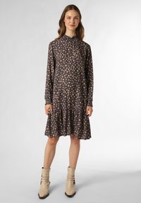 Langärmliges Kleid aus dunkelgrauem Stoff mit hellbeigen quadratischen Mustern, gerafftem Kragen und gestuftem Volant-Saum. Kombiniert mit beigen Ballerinas.